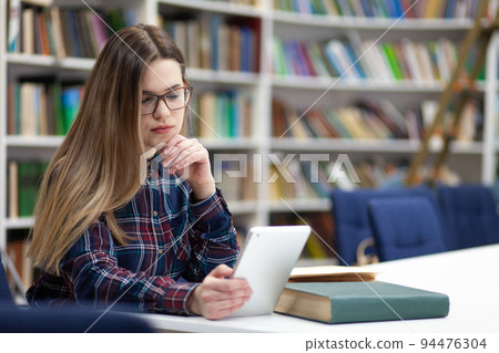 Girl in glasses working with tablet sitting in library. Student prefers to work with a tablet than books. Girl in glasses working with tablet sitting in library. Student prefers to work with a tablet than books. 94476304