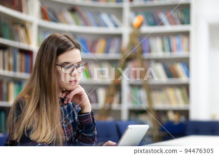 Girl in glasses working with tablet sitting in library. Student prefers to work with a tablet than books.. Girl in glasses working with tablet sitting in library. Student prefers to work with a tablet than books.. 94476305