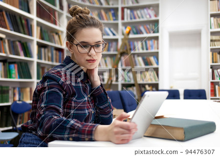 Girl in glasses working with tablet sitting in library. Student prefers to work with a tablet than books.. Girl in glasses working with tablet sitting in library. Student prefers to work with a tablet than books.. 94476307