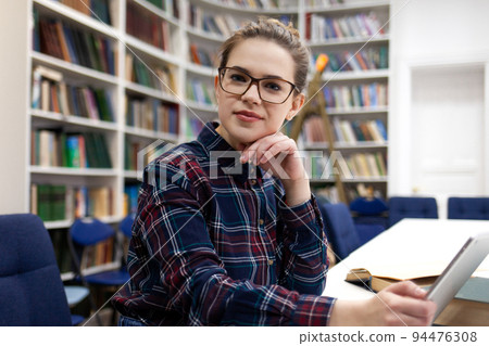 Girl in glasses working with tablet sitting in library. Student prefers to work with a tablet than books Girl in glasses working with tablet sitting in library. Student prefers to work with a tablet than books 94476308