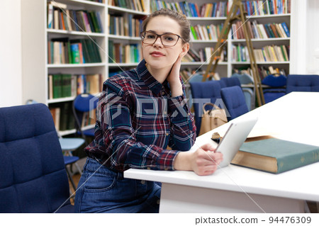 Girl in glasses working with tablet sitting in library. Student prefers to work with a tablet than books. 94476309