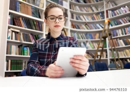 Girl in glasses working with tablet sitting in library. Student prefers to work with a tablet than books. Girl in glasses working with tablet sitting in library. Student prefers to work with a tablet than books. 94476310