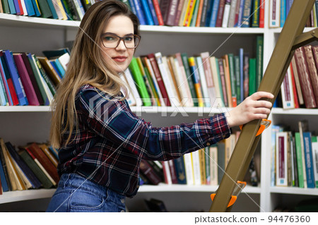Pretty student stands on a stepladder in the library to find the right book. Woman in public library.. Pretty student stands on a stepladder in the library to find the right book. Woman in public library.. 94476360