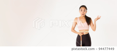 Fitness, healthy lifestyle and wellbeing concept. Surprised asian girl on diet, sportswoman wrap measuring tape around waist and look impressed as lose weight with workout 94477449