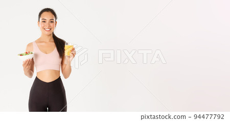 Sport, wellbeing and active lifestyle concept. Portrait of healthy and fit good-looking asian sportswoman holding orange juice and salad, morning breakfast before workout, white background Sport, wellbeing and active lifestyle concept. Portrait of healthy and fit good-looking asian sportswoman holding orange juice and salad, morning breakfast before workout, white background 94477792