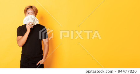 Portrait of lucky happy asian guy looking surprised and showing money, standing yellow background 94477820