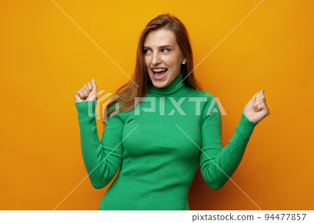 Portrait of an excited young woman celebrating success with raised arms against yellow background Portrait of an excited young woman celebrating success with raised arms against yellow background 94477857
