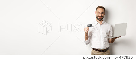 Online shopping. Handsome man showing credit card and using laptop to order in internet, standing over white background Online shopping. Handsome man showing credit card and using laptop to order in internet, standing over white background 94477939