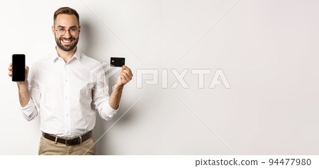 Business and online payment. Smiling handsome man showing mobile screen and credit card, standing over white background 94477980