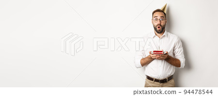 Holidays and celebration. Happy man having birthday party, making wish on b-day cake and smiling, standing against white background Holidays and celebration. Happy man having birthday party, making wish on b-day cake and smiling, standing against white background 94478544