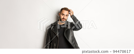 Handsome macho man in black biker jacket, touching his haircut and looking cool, white background 94478617