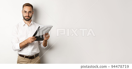 Serious employer looking at camera while reading documents on clipboard, having job interview, standing over white background Serious employer looking at camera while reading documents on clipboard, having job interview, standing over white background 94478719