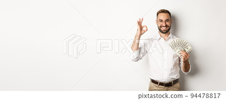 Satisfied young businessman showing money, make okay sign, earning cash, standing over white background Satisfied young businessman showing money, make okay sign, earning cash, standing over white background 94478817