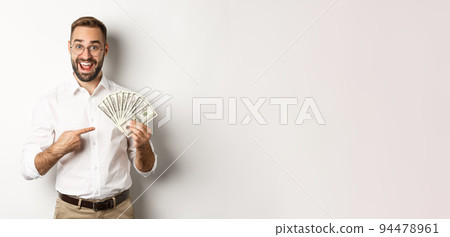 Excited businessman pointing at money, showing dollars and smiling, standing over white background 94478961