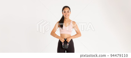 Sport, wellbeing and active lifestyle concept. Smiling strong and slim asian fitness girl, bodybuilding at home, holding workout equipment, do squats with kettlebell exercise, white background Sport, wellbeing and active lifestyle concept. Smiling strong and slim asian fitness girl, bodybuilding at home, holding workout equipment, do squats with kettlebell exercise, white background 94479110