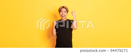 Portrait of triumphing confident asian man, fist pump and smiling happy, celebrating victory or achievement, standing yellow background 94479271
