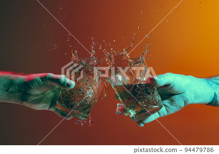 Male hands clinking glasses with whiskey over dark orange background in neon light Male hands clinking glasses with whiskey over dark orange background in neon light 94479786