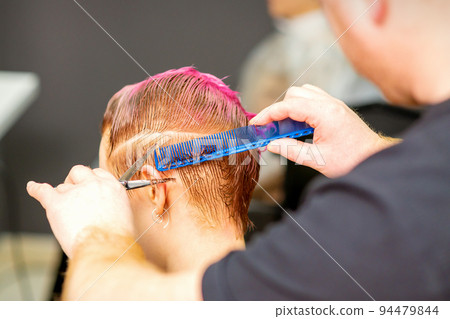 Haircut of dyed short pink wet hair of young caucasian woman by a male hairdresser in a barbershop Haircut of dyed short pink wet hair of young caucasian woman by a male hairdresser in a barbershop 94479844