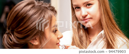 Makeup in the process. The makeup artist applies pink gloss lipstick on the lips of the beautiful face of the young caucasian woman in a beauty salon Makeup in the process. The makeup artist applies pink gloss lipstick on the lips of the beautiful face of the young caucasian woman in a beauty salon 94479903