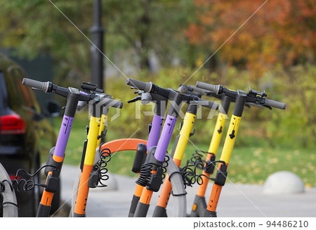 Several electric scooters for rent stand in one row in the parking lot in the autumn park. 94486210