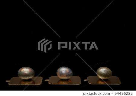 Three chicken eggs, painted in honor of the Easter holiday with dark paint, lie in one line on golden stands against a large black background - side view. 94486222