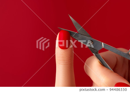 Manicured female hand with scissors on red background. Red matt nail design Manicured female hand with scissors on red background. Red matt nail design 94486330