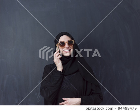 young modern muslim business woman using smartphone wearing sunglasses and hijab clothes in front of black chalkboard 94489479