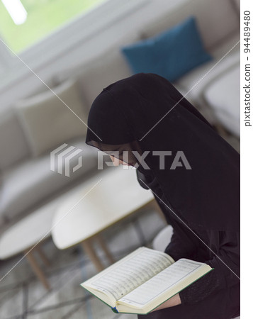 Young traditional muslim woman reading Quran on the sofa before iftar dinner during a ramadan feast at home 94489480