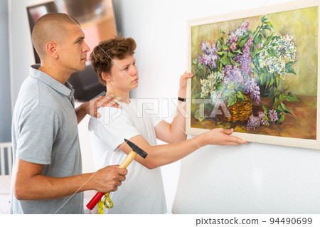 Father shows his son how to hang painting on the wall of house Father shows his son how to hang painting on the wall of house 94490699