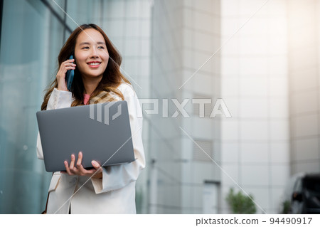 Asian businesswoman working on laptop and talking on cell phone at front building near office 94490917