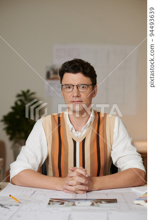 Portrait of serious confident architect in glasses sitting at desk with blueprints and looking at camera 94493639
