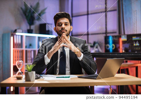 Indian bearded man in formal suit sitting at desk with modern laptop and looking at camera 94495194