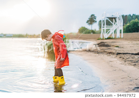Boy in a red raincoat and yellow rubber boots playing with water at the beach. School kid in a waterproof coat jumping in water at sea. Child having fun with waves at the shore 94497972