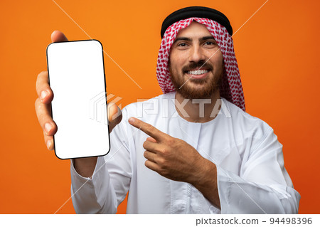 Young Arab muslim man in traditional clothes showing a mobile phone isolated on an orange background Young Arab muslim man in traditional clothes showing a mobile phone isolated on an orange background 94498396