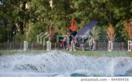 Wakeboarder making tricks while wakeboarding on lake. Young man surfer having fun wakesurfing in the cable park. Water sport, outdoor activity concept. 94499456