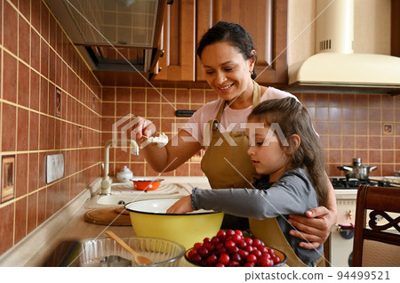 Beautiful African American young woman in chef apron, loving mom smiling and hugging her daughter, adorable multi-ethnic little child girl, while teaching her cooking homemade pie in the home kitchen Beautiful African American young woman in chef apron, loving mom smiling and hugging her daughter, adorable multi-ethnic little child girl, while teaching her cooking homemade pie in the home kitchen 94499521