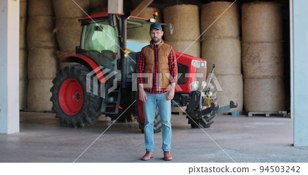 Handsome farmer standing in a shirt and orange vest smiling at the camera, on a tractor and haystack background. Cheerful male worker in agricultural farm. Machine for agriculture. 94503242