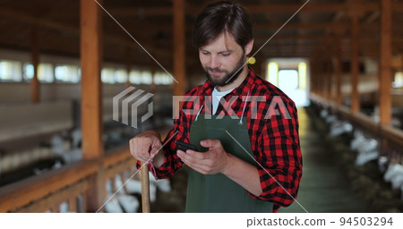 Handsome male farmer in green apron in goat stable and texting message on smartphone. Man farmer tapping and scrolling on mobile phone. Modern livestock farming business. 94503294