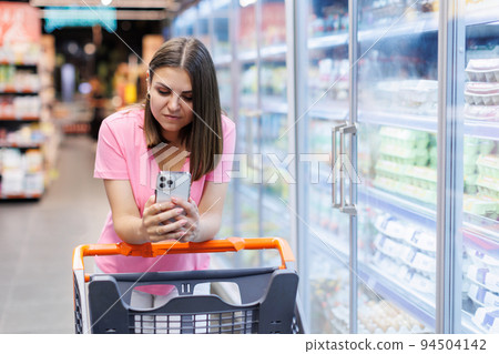 Happy caucasian woman using mobile phone in grocery shopping. Checklist application buying food products in supermarket. 94504142