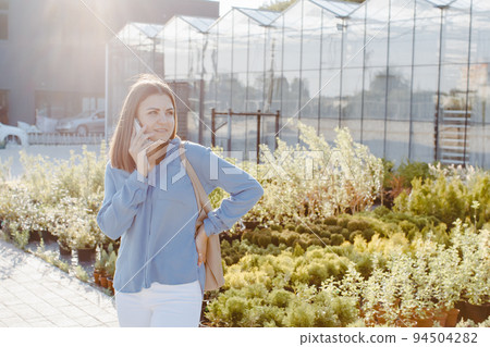 A young pretty girl, dressed in shirt and jeans, stands near the flower center and talking to the phone. The woman is going to buy green plants for decoration. Landscaping concept A young pretty girl, dressed in shirt and jeans, stands near the flower center and talking to the phone. The woman is going to buy green plants for decoration. Landscaping concept 94504282