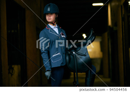 Portrait of young girl, jockey and horsewoman in competition uniform and helmet standing stands in stable before the start of horse race Portrait of young girl, jockey and horsewoman in competition uniform and helmet standing stands in stable before the start of horse race 94504456