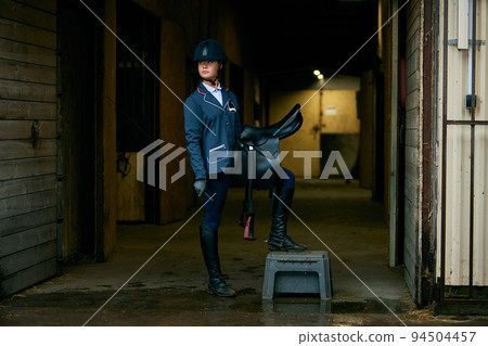 Portrait of young girl, jockey and horsewoman in competition uniform and helmet standing stands in stable before the start of horse race Portrait of young girl, jockey and horsewoman in competition uniform and helmet standing stands in stable before the start of horse race 94504457