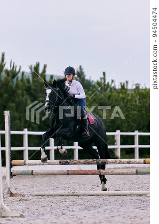 Jump over barrier. Young sportive girl, teen training at riding arena in summer day, outdoors. Dressage of horses. Horseback riding. Model in sports uniform Jump over barrier. Young sportive girl, teen training at riding arena in summer day, outdoors. Dressage of horses. Horseback riding. Model in sports uniform 94504474