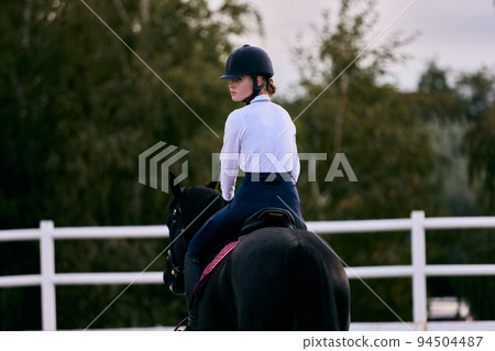Sports training. Young sportive girl, professional rider in sports uniform and helmet practicing at riding arena in summer day, outdoors. Horseback riding 94504487