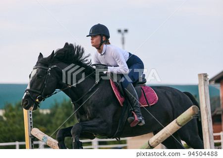 Sports training. Young sportive girl, professional rider in sports uniform and helmet practicing at riding arena in summer day, outdoors. Horseback riding 94504488