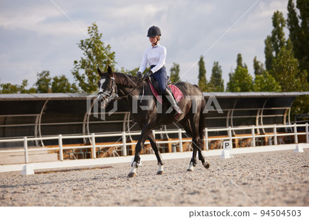 Equestrian sport. Portrait of teen girl, beginner female rider training at riding arena in autumn day, outdoors. Dressage of horses. Horseback riding 94504503