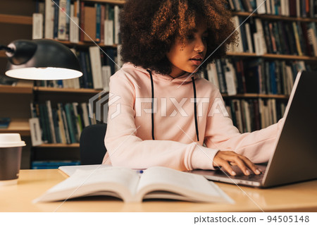 Girl with curly hair typing on laptop in library. High school student preparing for exams at evening.	 94505148