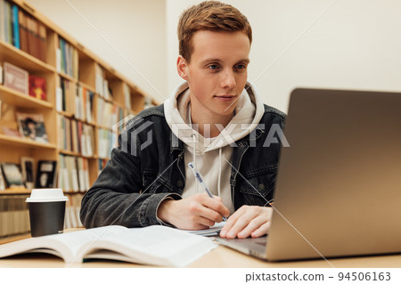 Male student writing and looking on laptop. Guy in casuals preparing exams at desk in library.	 94506163