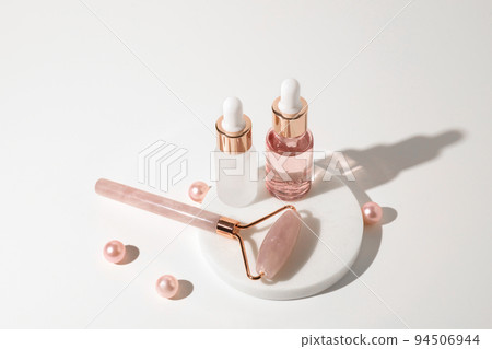 Massage quartz roller and two glass bottles of cosmetic liquid transparent gel on white background with pink pearls. Facial massage and relaxation. Gua Sha massage tools. Massage quartz roller and two glass bottles of cosmetic liquid transparent gel on white background with pink pearls. Facial massage and relaxation. Gua Sha massage tools. 94506944