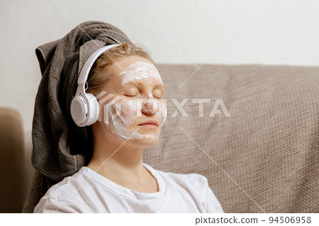 Young woman with casual clothes, towel on head and beauty mask on face sitting on couch at home, resting and listening music. Cosmetic for women, skin care. Self care, time for yourself. Relax. 94506958